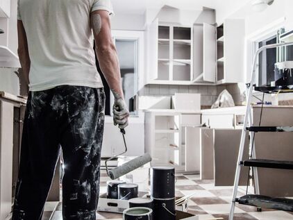 Man holding paint roller and covered in white paint and surrounded by freshly painted open kitchen cabinets.